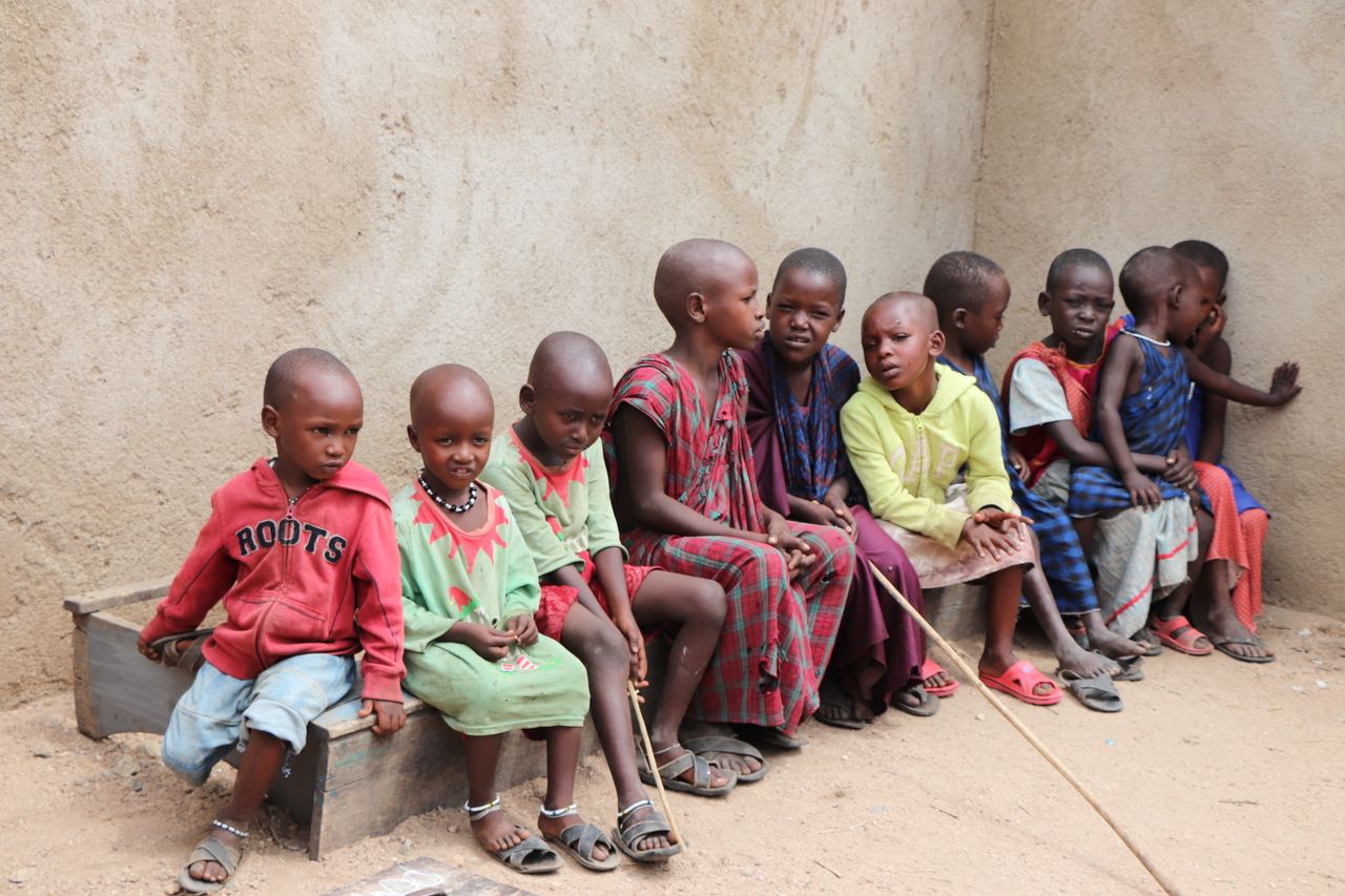 Children in colorful clothing sitting together on a wooden bench outside a concrete wall.