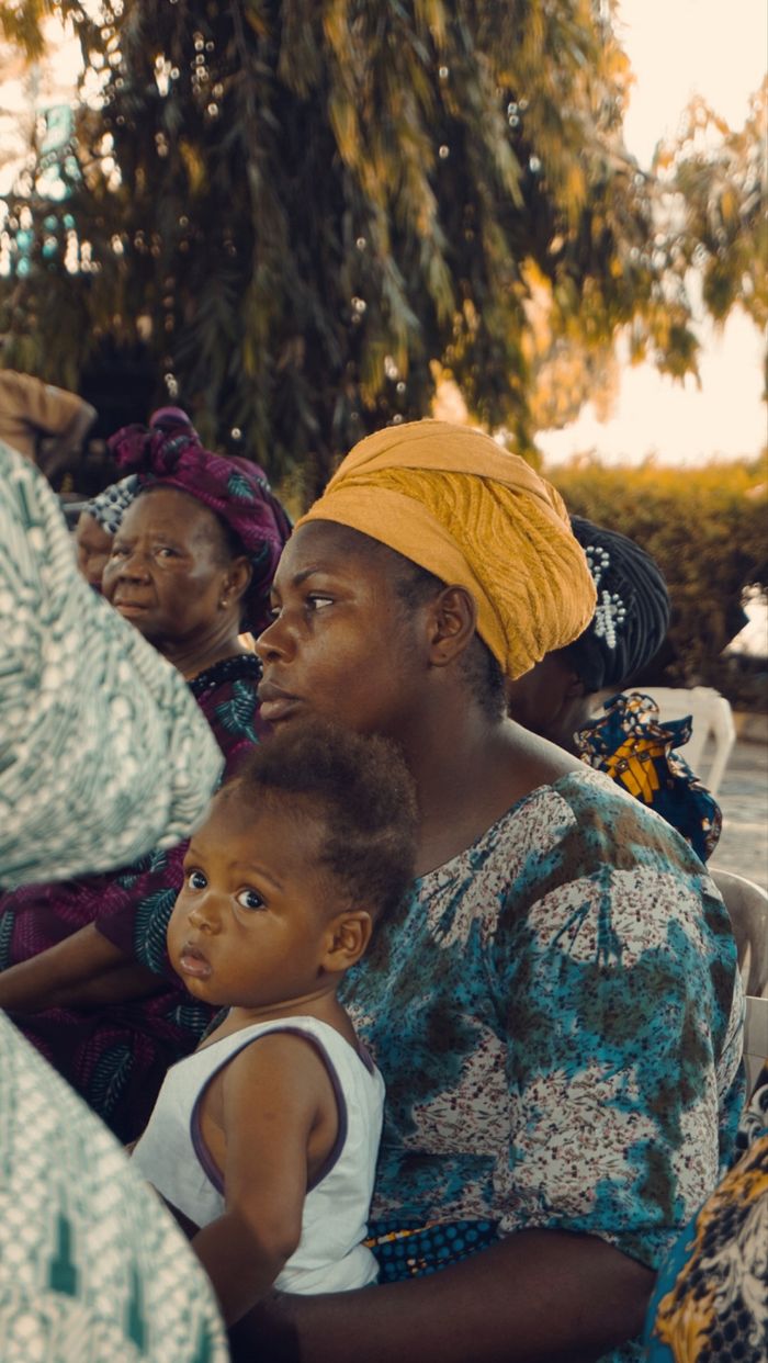 A mother in a yellow turban holds her child during an outdoor gathering, capturing a serene moment.